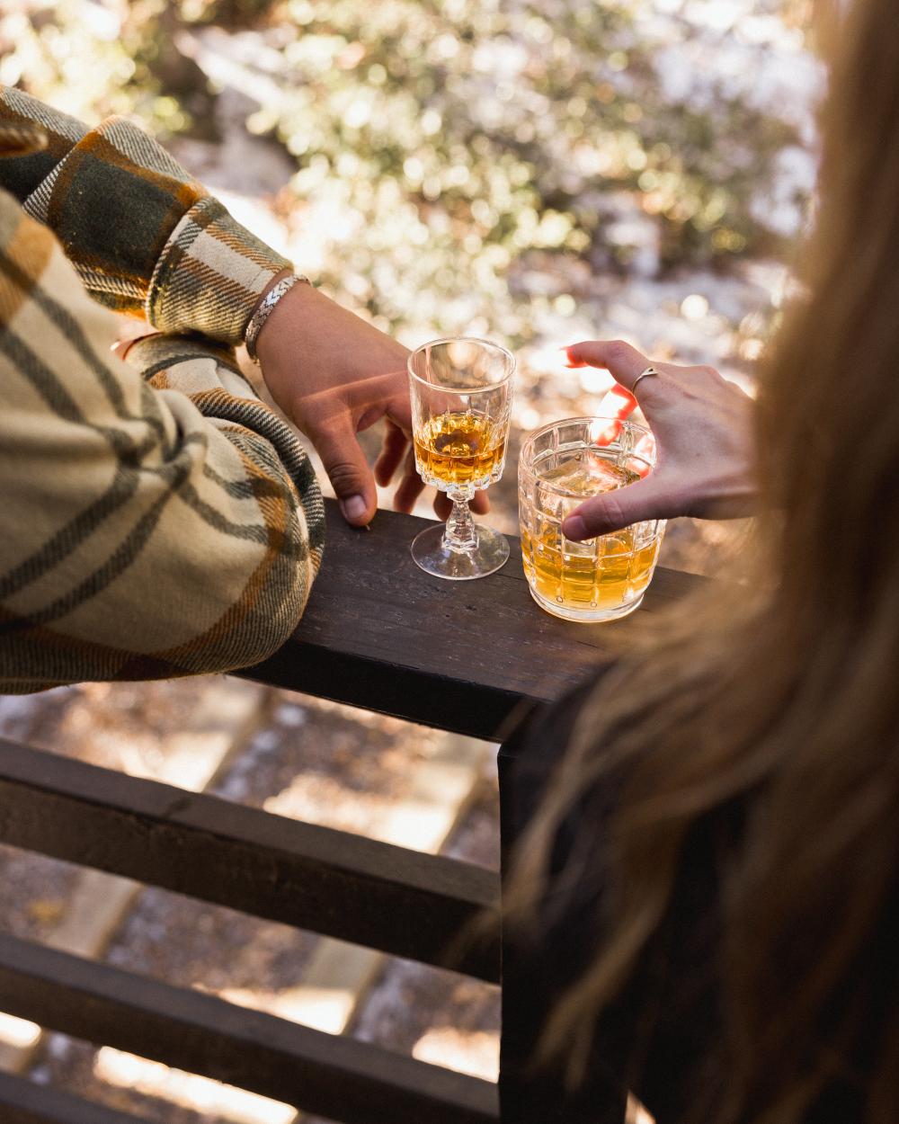 two people holding glasses of whiskey on a railing